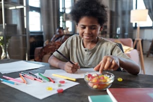 Glücklicher afrikanischer Schuljunge, der Süßigkeiten aus der Schüssel nimmt, während er am Tisch sitzt und ein Bild gegen seine Mutter malt