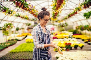 Florista bonito sorridente com cabelos castanhos, avental e óculos colocando luvas de jardinagem enquanto está em pé na estufa.
