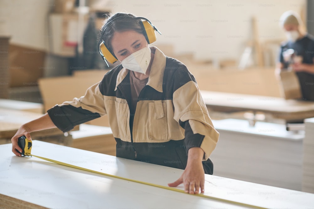 Young woman in protective headphones and mask using tape measure to make measurements of wooden board
