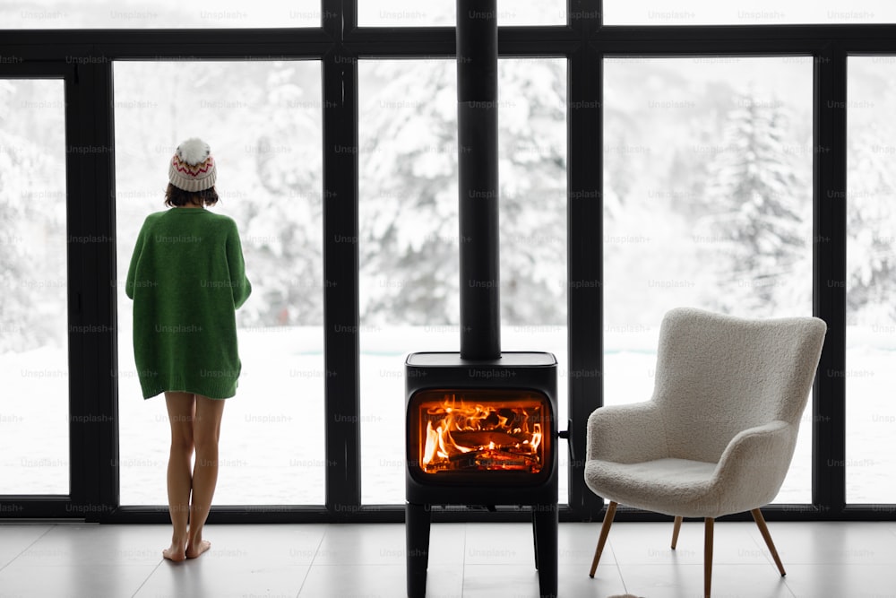 Woman standing with cup by the fireplace near the window at modern house on nature during winter time. Concept of winter mood and comfort at home. Idea of recreation in cabins on mountains