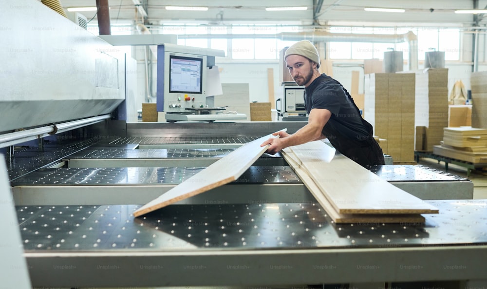 Jeune ouvrier manuel mettant des planches de bois coupées en pile tout en utilisant une machine à l’usine