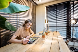 Mujer creativa en traje doméstico cómodo que trabaja en tableta digital en la oficina en casa acogedora y con estilo