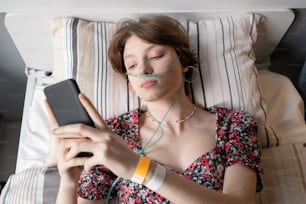 High angle view of sick teenage girl breathing with oxygen tube while lying on the bed and talking online with friends using her mobile phone