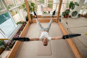 スポーツセンターでの個人的な空中ヨガのトレーニング中にロープに固定された彼の腕と脚で床にぶら下がっているアクティブウェアの若い男性