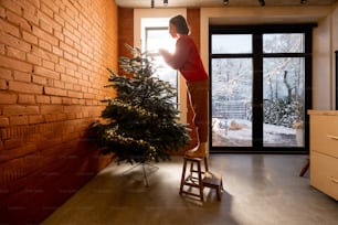 Mujer joven decorando el árbol de Navidad en casa. Concepto de confort en el hogar y preparación para unas vacaciones de invierno. Muchacha con suéter rojo, sala de estar con ventanas que dan al jardín nevado