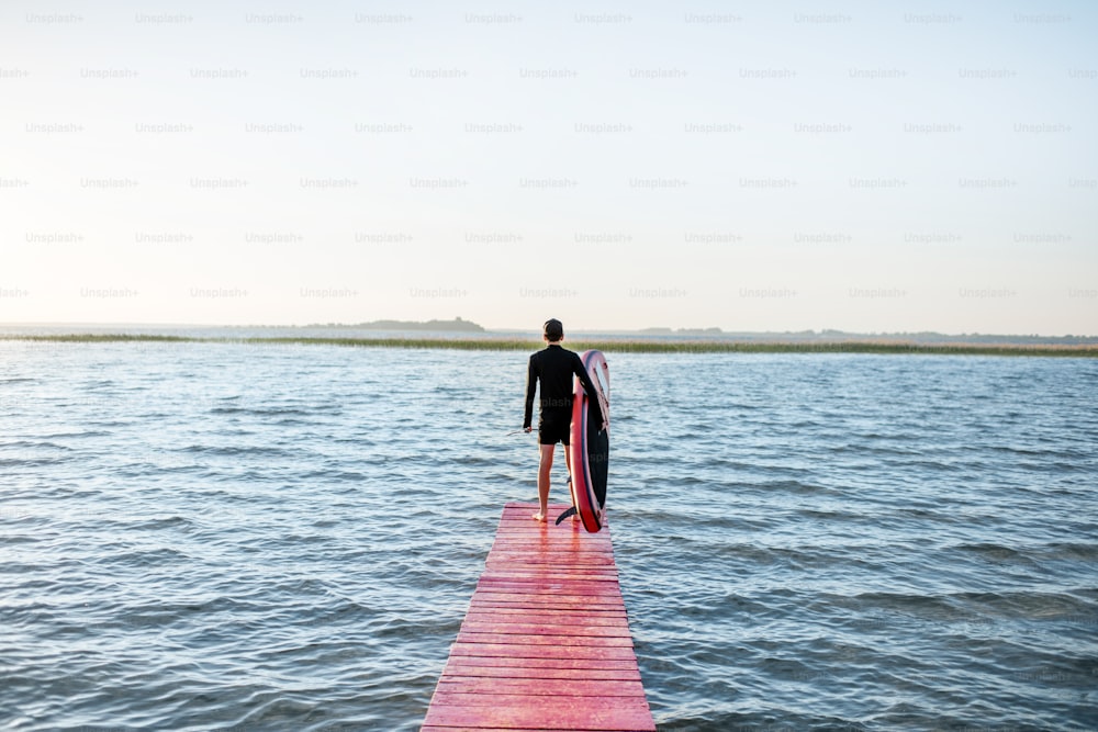 日の出の間に桟橋にパドルボードを持って立っている男性と湖の風景