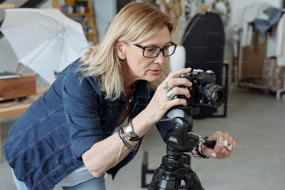 Fotografo maturo concentrato in occhiali che guarda attraverso la fotocamera sul treppiede mentre fa un servizio fotografico