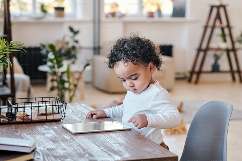Concentrated modern mixed race toddler with curly hair standing at desk and using tablet in living room