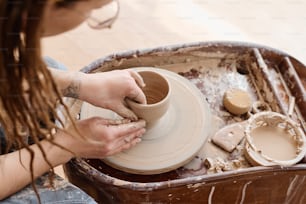 View from above of young creative female pitcher working over new clay item while sitting in front of rotating pottery wheel in workshop