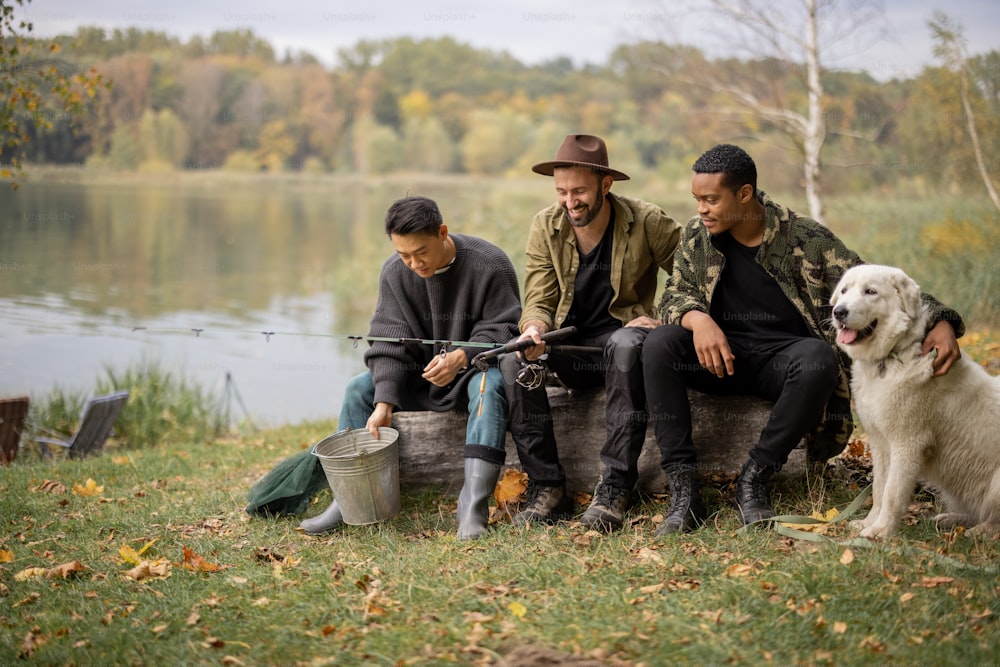 Multiracial male friends resting and talking in nature. Men after fishing on river or lake coast. Concept of leisure and weekend in nature. Idea of friendship and spending time together with dog
