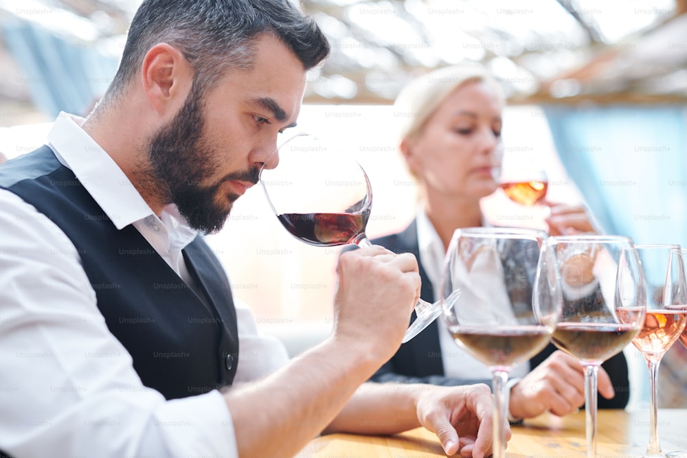 Young serious sommelier feeling smell of red wine while holding wineglass by his nose during work in cellar
