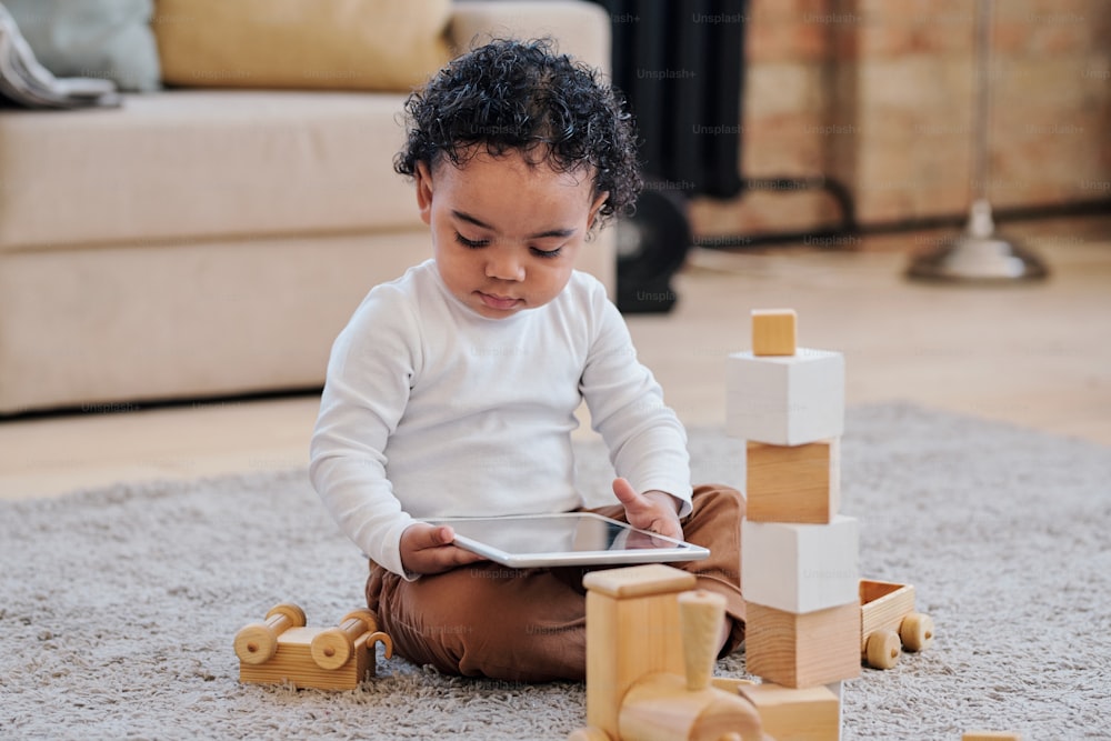Serious curious African-American toddler boy in white shirt sitting on floor among wooden toys and using tablet