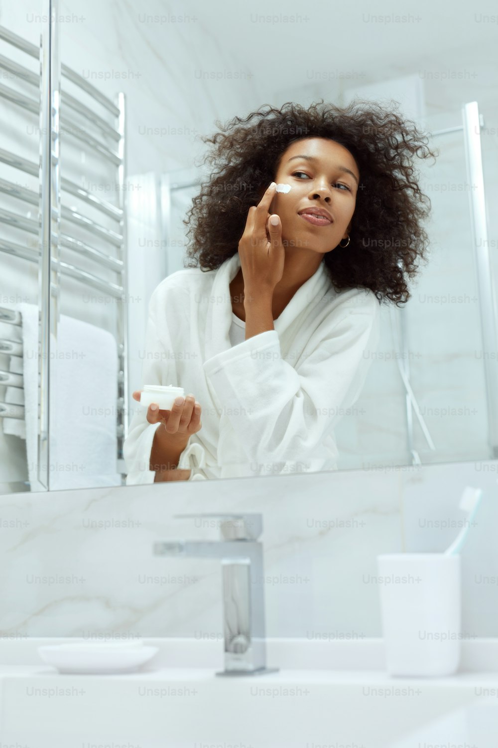 Skin care. Woman applying cosmetic cream on face looking in mirror at bathroom. Portrait of african girl with natural makeup, clean facial skin applying moisturizing lotion
