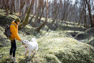 Young woman in hiking clothes and backpack spend time together with big white dog in green spring forest. Enjoys and explore of tranquil nature. Copy space.
