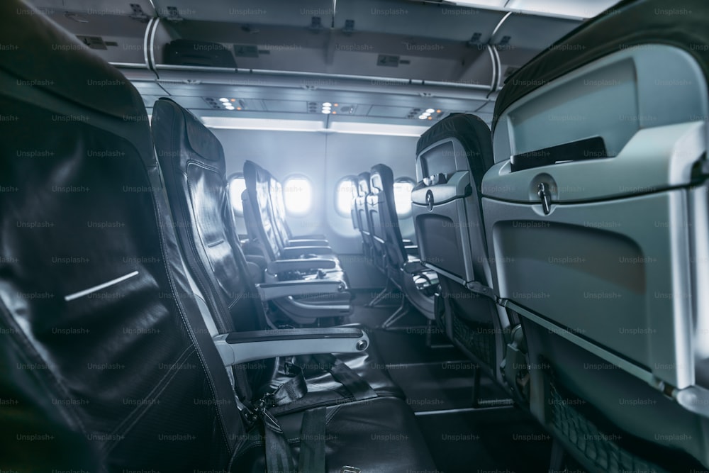 A dark aircraft interior: the row of modern empty thin leather seats with armrests down, shallow depth of field, selective focus in the foreground, opened luggage shelves; wide-angle