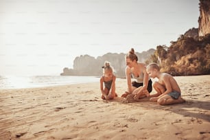 Smiling Mom and her two cute children playing together in the sand during summer vacation at the beach