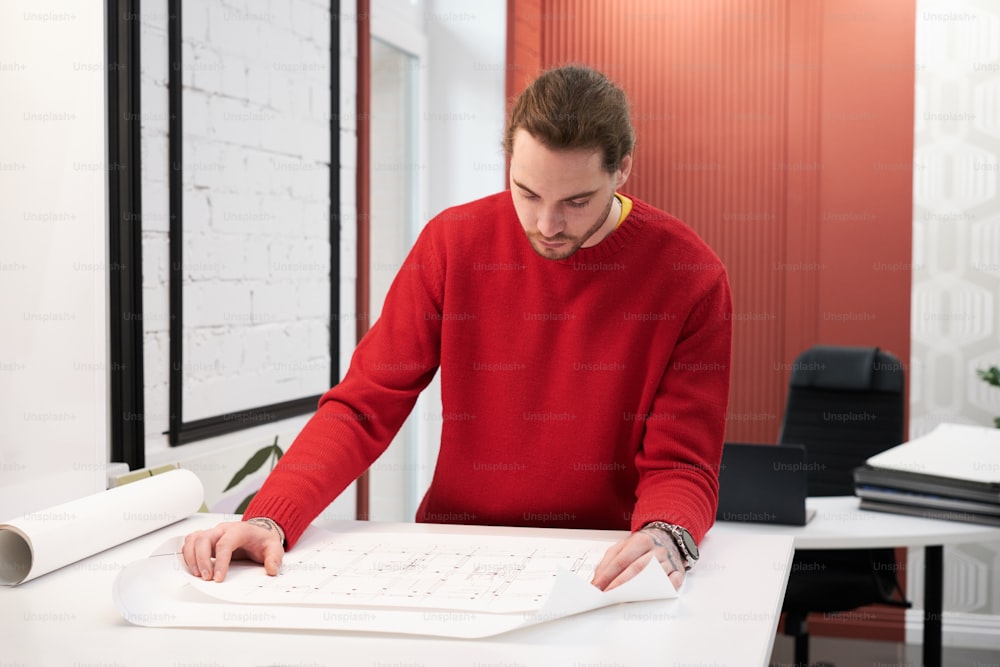 Young architect examining blueprint of new building at table during his work at office