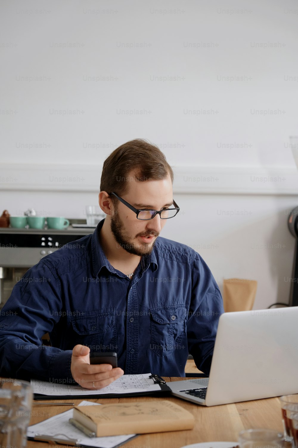 Ritratto di uomo serio in occhiali e camicia blu che lavora su laptop e smartphone mentre si siede al tavolo di legno contro la parete bianca
