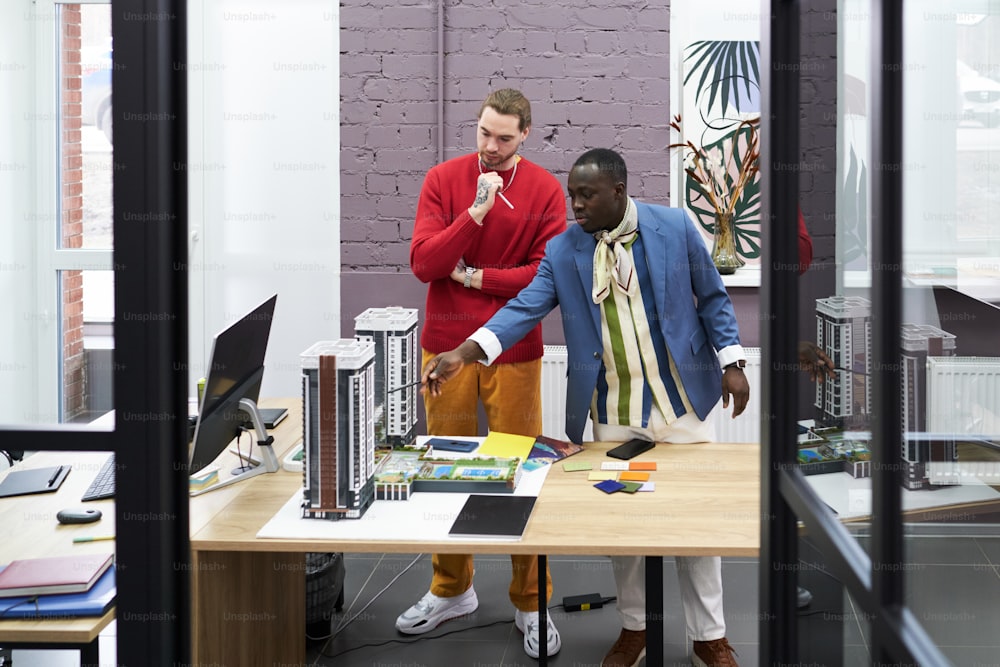 African young architect pointing at mock-up of buildings on table and talking to his colleague