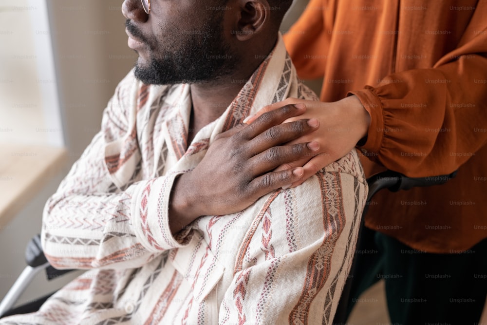 Junger Afroamerikaner im Rollstuhl bedeckt die Hand einer weiblichen Pflegekraft auf seiner Schulter, während er durch das Fenster schaut