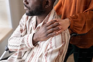 Jeune homme afro-américain en fauteuil roulant couvrant la main d’une soignante sur son épaule tout en regardant par la fenêtre