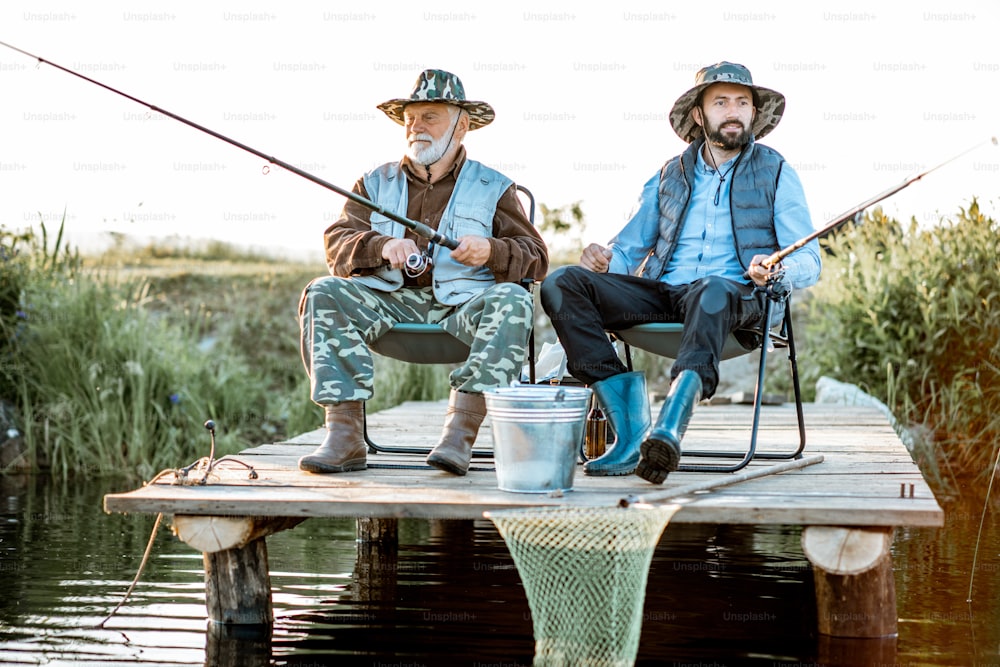 Grandfather with adult son fishing together on the wooden pier during the morning light. View from the side of the lake