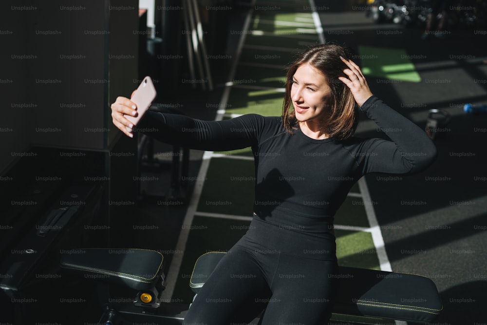 Retrato de la mujer morena joven descansando en el deporte ropa activa tomando selfie en el club de fitness