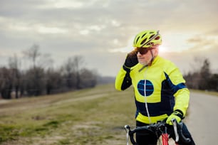 An old bicyclist talking on the phone while taking a break in nature.