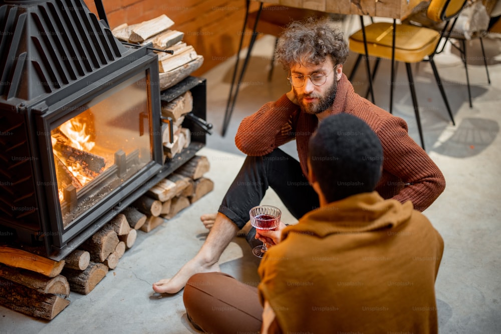 Dois homens conversando enquanto estão sentados juntos junto à lareira em chamas em uma casa aconchegante. Conceito de relações homossexuais e aconchego no inverno. Ideia de famílias gays multinacionais