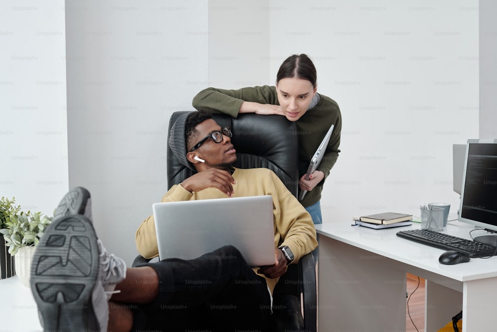 Two young intercultural programmers in casualwear consulting at meeting while black man with laptop sitting in armchair