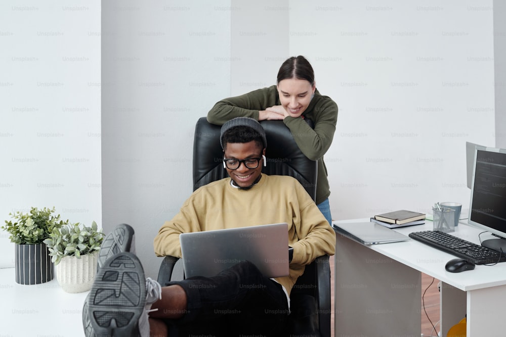 Due giovani sviluppatori di software multirazziale felici che guardano lo schermo del computer portatile sulle ginocchia dell'uomo afroamericano seduto in poltrona
