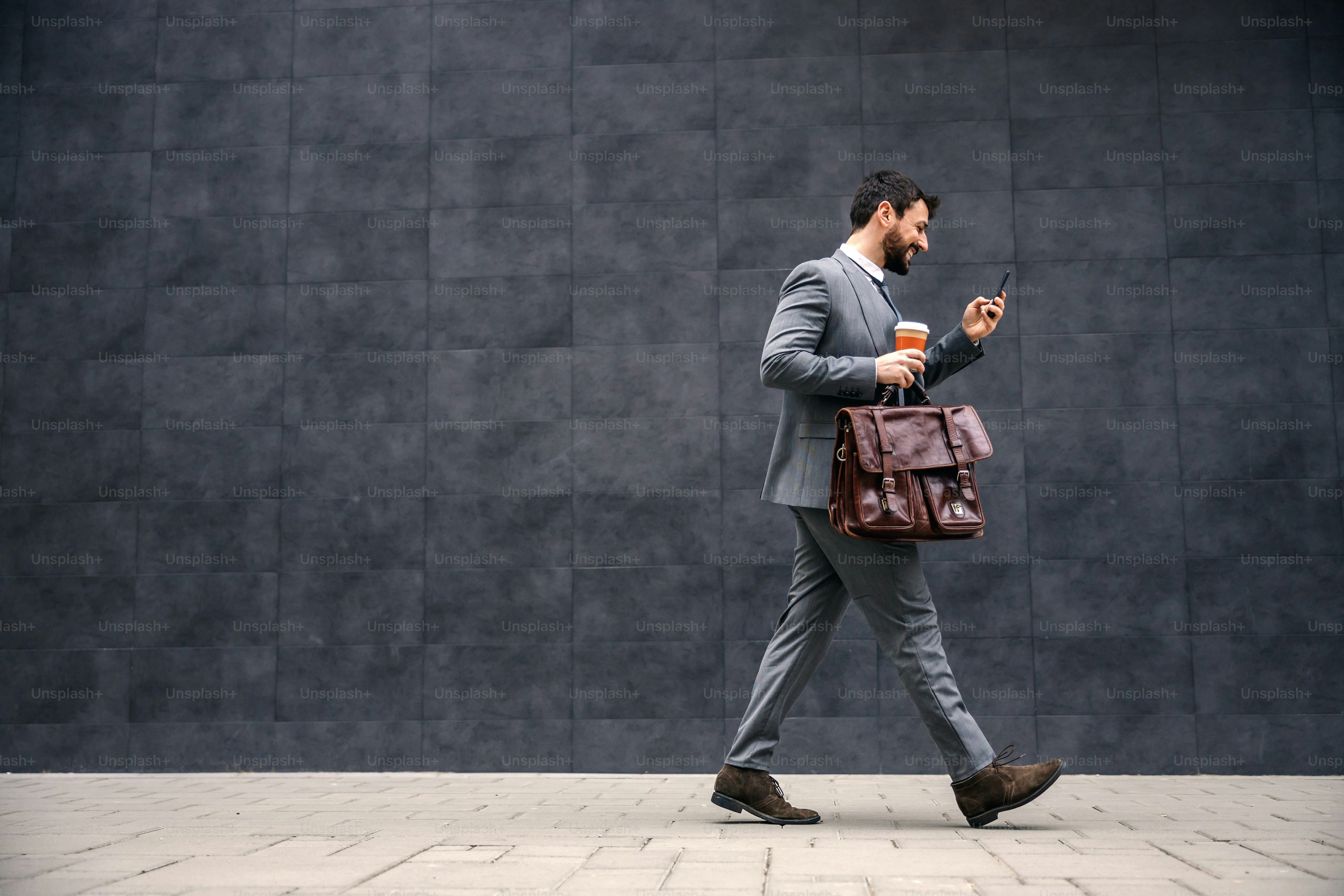 Young attractive bearded businessman walking on the street in the morning, reading message on smart phone and going to work