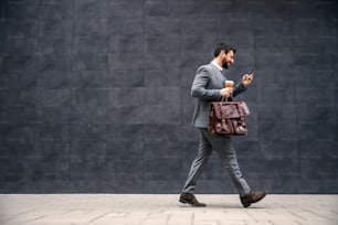 Giovane uomo d'affari barbuto attraente che cammina per strada al mattino, legge un messaggio sullo smartphone e va al lavoro.