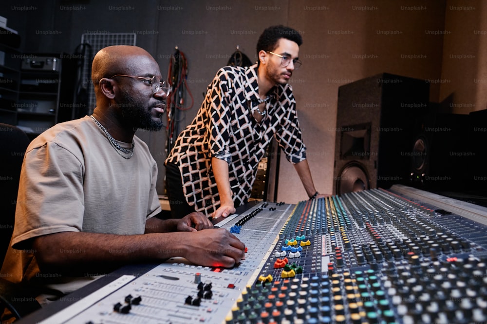 Engenheiro de som negro maduro moderno e jovem cantor elegante trabalhando em faixa de música em estúdio de gravação