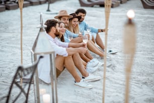 Group of young friends hanging out together with wine glasses, having a festive meeting and enjoying evening time on the sandy beach