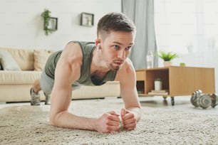 Young active man with earphones doing plank exercise on the carpet in living-room while training at home during period of quarantine