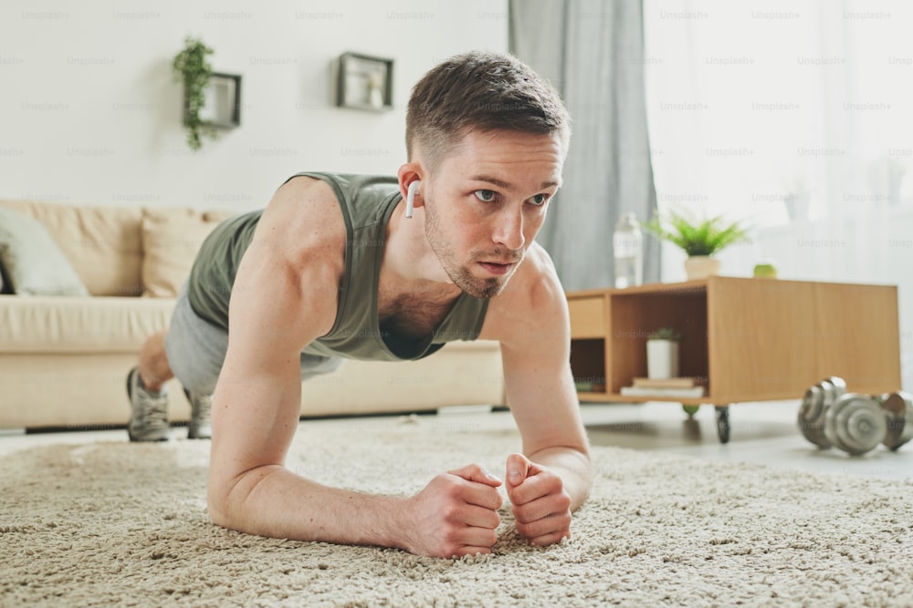 Young active man with earphones doing plank exercise on the carpet in living-room while training at home during period of quarantine