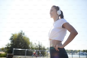 Smiling optimistic young lady in wired headphones standing outdoors and looking into distance while thinking about new achievements
