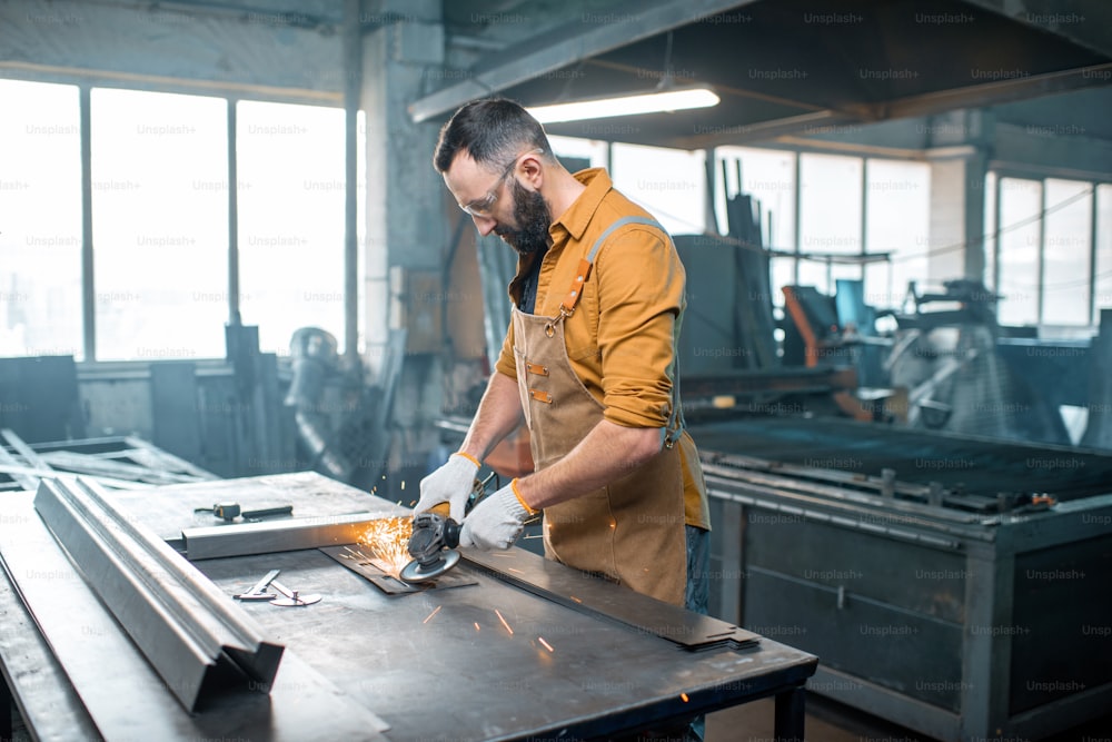エプロンを着た金属産業の労働者が、工場で鉄鋼製品を研削する金属加工をしています。金属製品の製造におけるハンサムな職人。高品質の写真