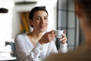 Young smiling brunette woman with cup of tea or coffee enjoying the drink during conversation with her friend in cafe