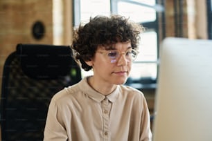 Young employee in eyeglasses with curly hair looking at computer monitor during her work at office