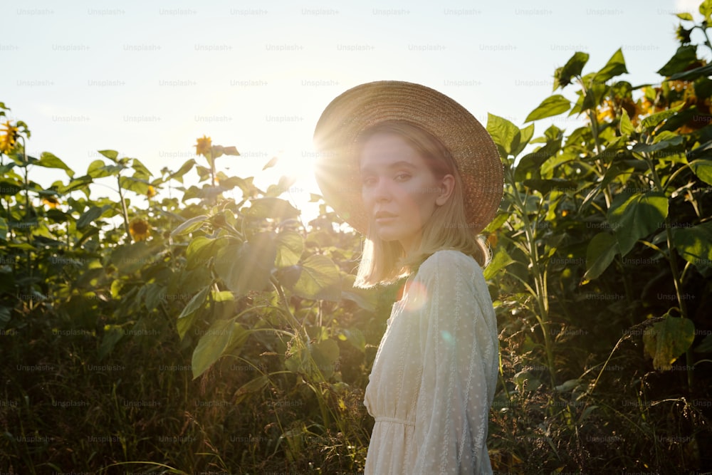 麦わら帽子と白いドレスを着た美しい若いブロンドの女性が、日の出にひまわりを背景にカメラの前に立っている間、あなたを見ています