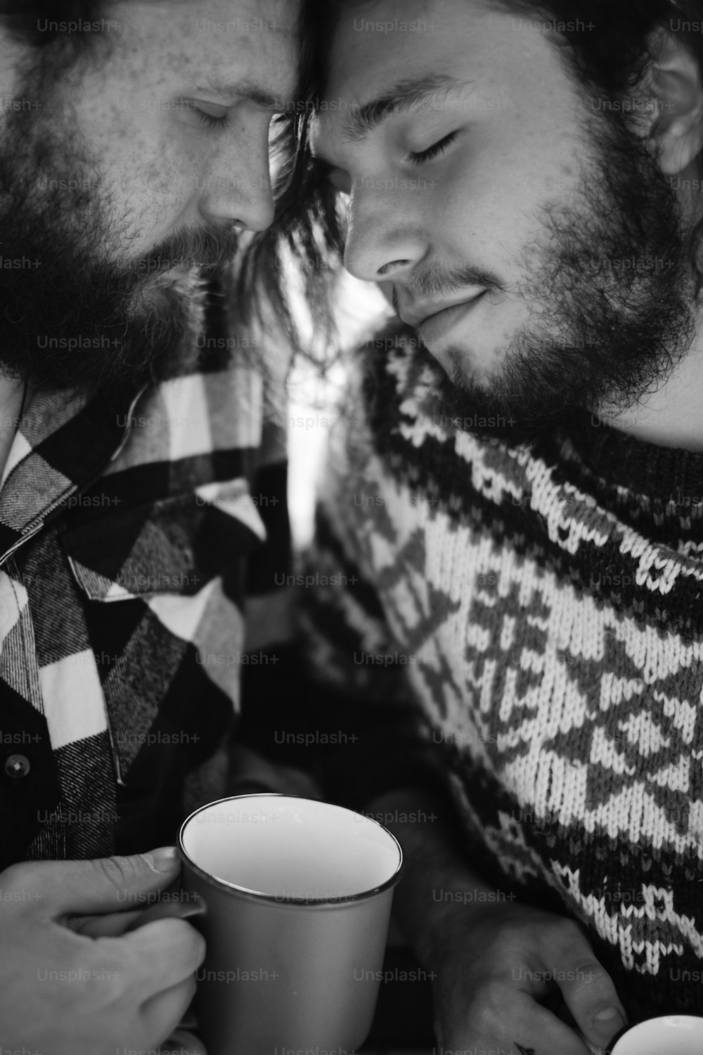 Black and white photo portrait of two bearded men with long hair leaning on each other, they are wearing warm sweater and plaid shirt and holding cups in hands
