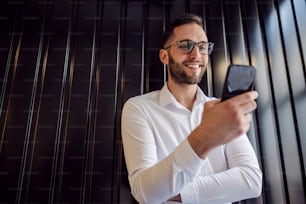 Low angle view of young happy geeky man leaning on the wall and using smart phone for checking on messages on social media.