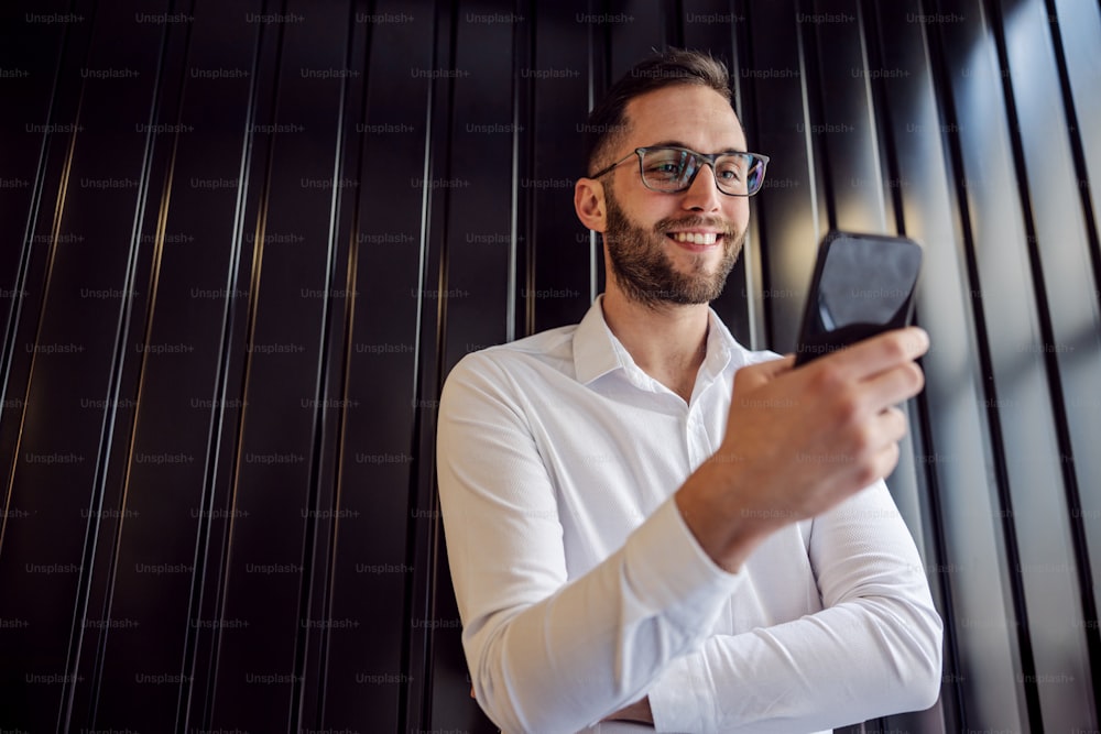 Low angle view of young happy geeky man leaning on the wall and using smart phone for checking on messages on social media.