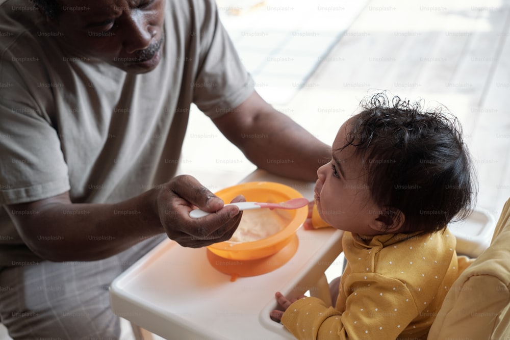 High angle view shot of mature African American man sitting in front of his baby daughter feeding her with puree