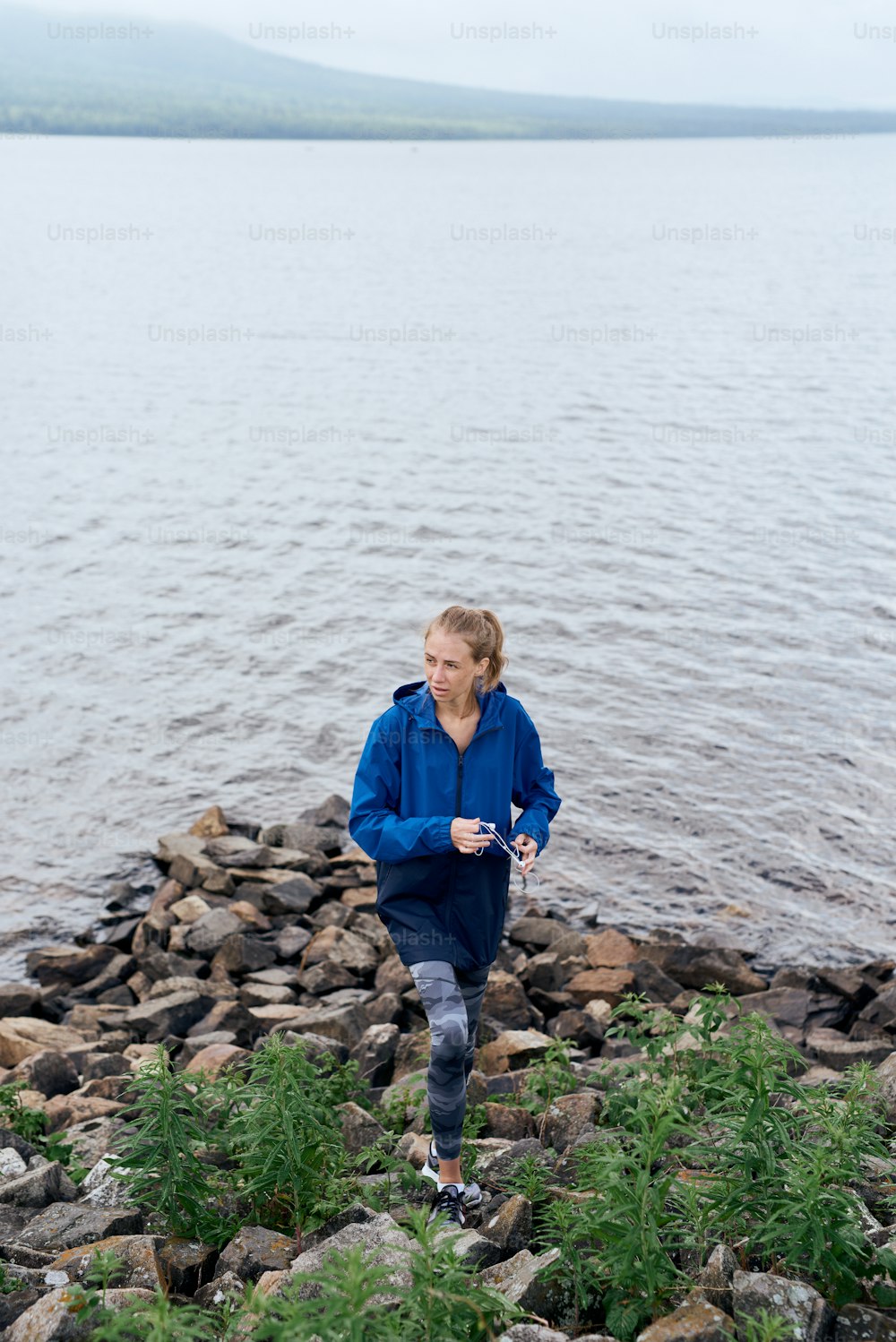 Full length of skinny woman standing on rocky river bank outdoor, listening to music in earphones, wearing blue raincoat