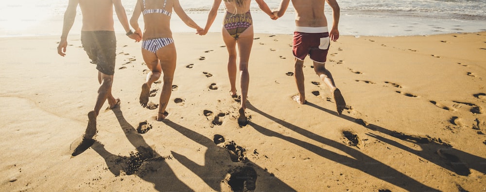 Vista de volta as pessoas nas férias de verão correndo na areia da praia para o mar de água todos juntos em amizade de mãos dadas - biquíni grupo homens e mulheres se divertem no pôr do sol com sombras