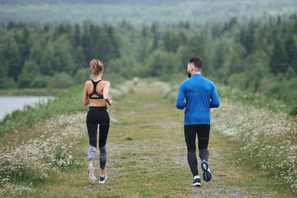 Two mid age man and woman jogging outdoor, warming up muscules, in summer, on gloomy day, at the road with perspective and scenic view, full length, from the back, running away