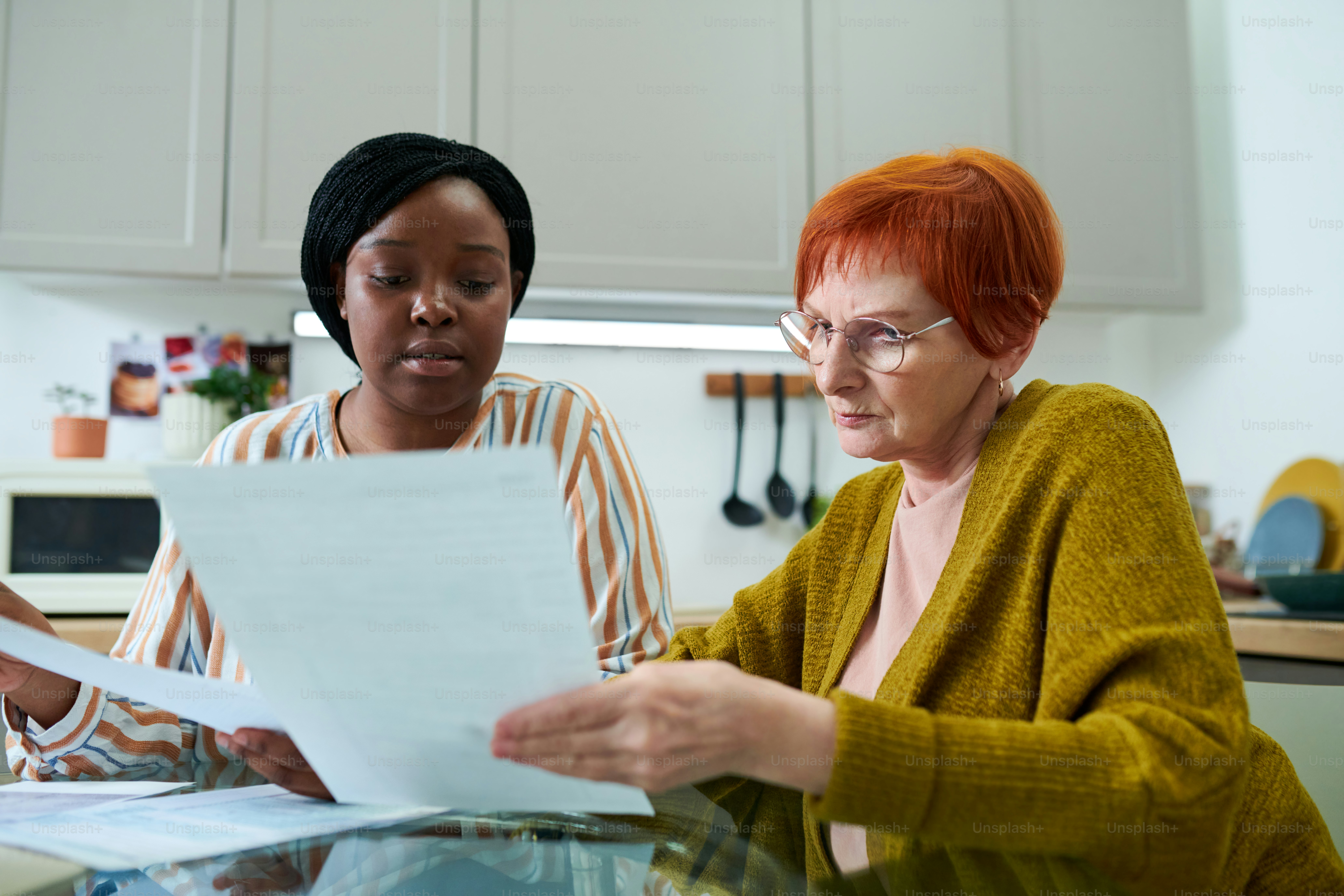 African female volunteer helping elderly woman to pay bills, they sitting at table in kitchen and examining documents